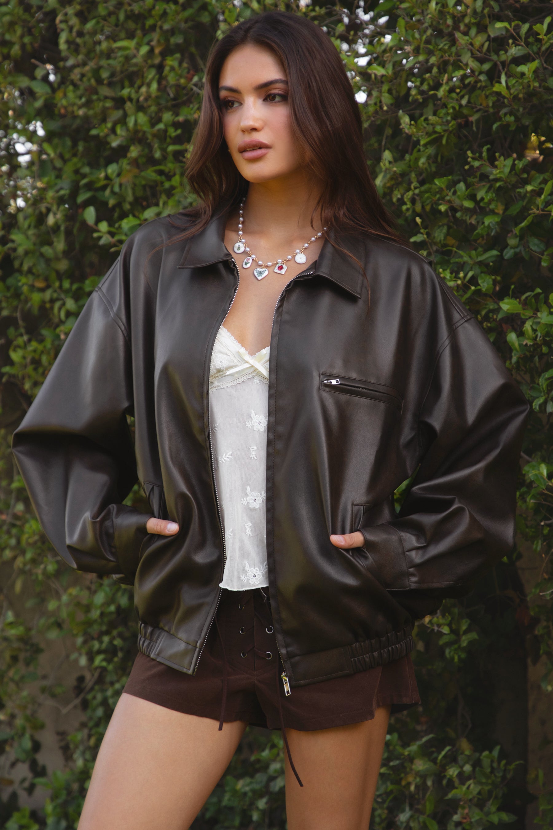 Women wearing TIC TOC GIANA Oversized PU Leather Bomber Jacket in dark brown with collared neckline, puff sleeves, and elastic waistband over a white baby doll floral top, suede lace up shorts, and brown frye boots standing against a green leafy background. 