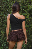 Person wearing a black top and layered brown bloomer shorts against a green leafy background