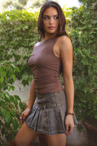 Woman wearing a brown top and denim skirt standing outdoors with greenery in the background.