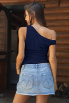 Woman wearing a navy one-shoulder top and denim skirt in front of a wooden wall.