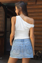 Woman wearing a white one-shoulder top and denim skirt in front of a wooden wall.