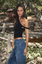 Women wearing TIC TOC SHANTAL One Shoulder Top in black with twisted strap, ruched seams, and studded torso detail paired with a denim blue jeans. 
