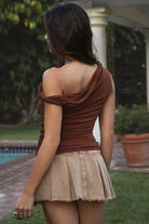 Women wearing TIC TOC SHANTAL One Shoulder Top in copper with twisted strap, ruched seams, and studded torso detail paired with a denim brown pleated mini skirt. 