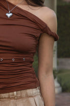 Women wearing TIC TOC SHANTAL One Shoulder Top in copper with twisted strap, ruched seams, and studded torso detail paired with a denim brown pleated mini skirt. 