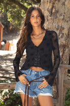 Women wearing a TIC TOC VIVIANA Floral Flocked Mesh Long Sleeve Top in black featuring a low V neckline, sheer sleeves, and asymmetrical cropped hem with lettuce trim paired with blue denim shorts in an outdoor setting