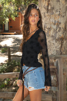 Women wearing a TIC TOC VIVIANA Floral Flocked Mesh Long Sleeve Top in black featuring a low V neckline, sheer sleeves, and asymmetrical cropped hem with lettuce trim paired with blue denim shorts in an outdoor setting