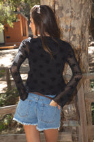 Women wearing a TIC TOC VIVIANA Floral Flocked Mesh Long Sleeve Top in black featuring a low V neckline, sheer sleeves, and asymmetrical cropped hem with lettuce trim paired with blue denim shorts in an outdoor setting