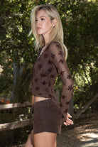 Women wearing a TIC TOC VIVIANA Floral Flocked Mesh Long Sleeve Top in brown featuring a low V neckline, sheer sleeves, and asymmetrical cropped hem with lettuce trim paired with brown suede shorts in an outdoor setting