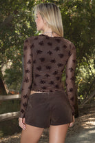 Women wearing a TIC TOC VIVIANA Floral Flocked Mesh Long Sleeve Top in brown featuring a low V neckline, sheer sleeves, and asymmetrical cropped hem with lettuce trim paired with brown suede shorts in an outdoor setting