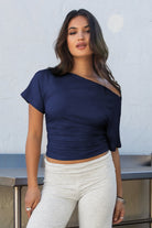 Woman model wearing TIC TOC TH31574BA RAYAE heather white asymmetric off-the-shoulder top with draped mid-sleeves and soft, brushed stretch fabric standing against a light-colored wall