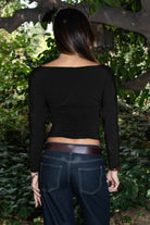 Woman wearing TIC TOC TH31575 MARCELLE brown asymmetric off the shoulder long sleeve top with wide boat neckline and ruched waist paired with dark denim standing in front of green foliage.