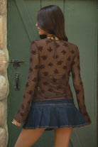 Woman wearing TIC TOC LINETTE Floral Flocked Mesh Long Sleeve Top in brown with high mock neckline, bell sleeves, and button front detail and blue denim skirt standing against a stone wall and green door.