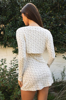 Woman wearing TIC TOC TH31985-LYNN-CC LYNN floral pointelle print long sleeve bolero top featuring a wide boat neckline with front cutout, single front buckle closure, long fitted sleeves, cropped bolero length, and ribbed floral pointelle fabric with a soft airy stretch figure-hugging fit, styled layered over a matching floral pointelle cami and lounge pants for a feminine cozy set.