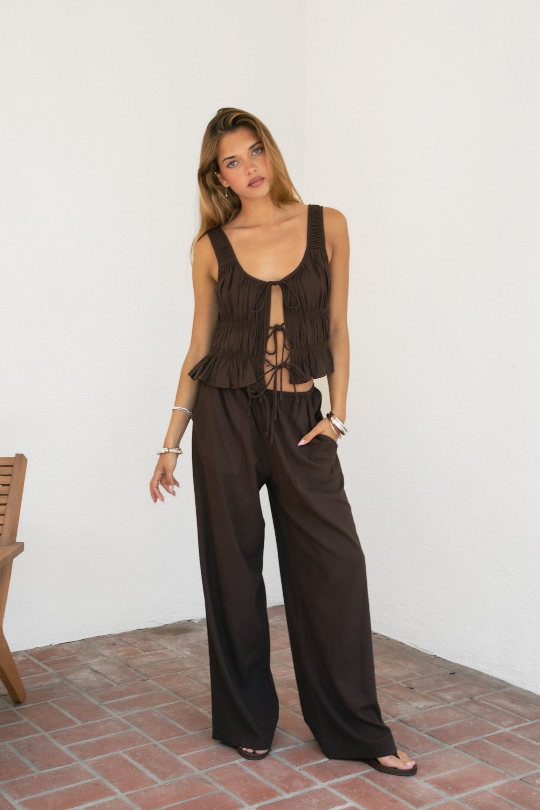 Woman wearing SOLSTICE ruched tie front round neck cami top featuring soft ruched texture throughout the body, three self-tie front closures, deep round neckline, wide shoulder straps, and a lightweight viscose linen blend with ruffled hem detail for relaxed resort styling.