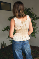 Woman wearing LENA smocked ribbon tie peplum tank top featuring a romantic scoop neckline with scalloped lace trim, front ribbon tie and button detail, smocked waist, wide shoulder straps with satin ribbon loop accents, and soft lightweight lyocell fabric.