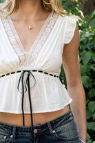 Woman wearing REVENE deep V-neck lace trim cropped blouse top featuring lace trim along the front and back neckline, ruffle cap sleeves, contrast ribbon underbust detail, front tie bow closure, elastic underbust seam with gathered peplum hem, and lightweight gauze cotton fabric.