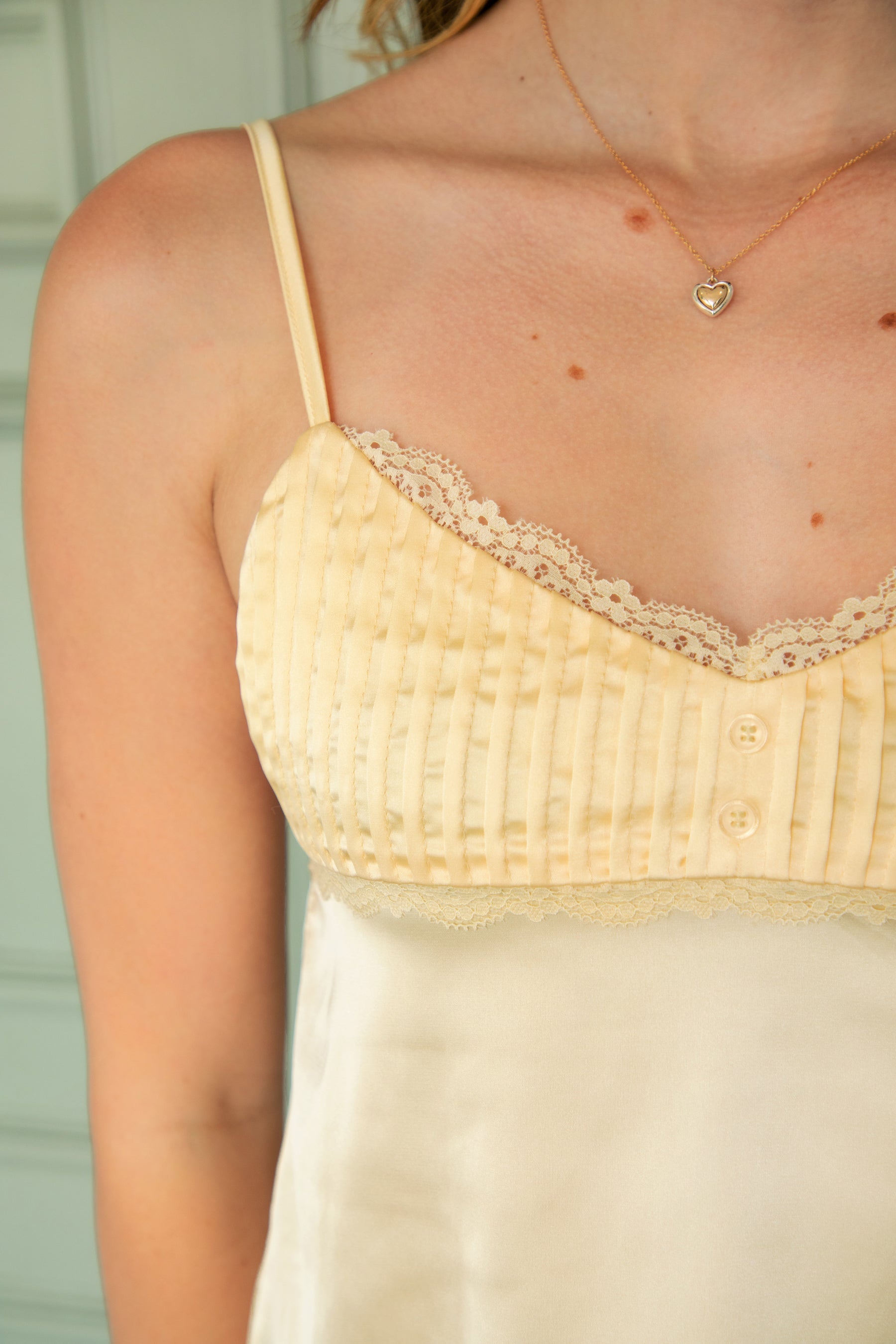 Woman wearing BUTTERCUP satin lace trim cami top featuring soft stretch satin fabric, delicate lace trim along the neckline and bust, ruched bust detail with decorative front button accent, adjustable spaghetti straps, and a self tie open back for feminine summer styling.