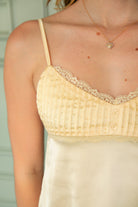 Woman wearing BUTTERCUP satin lace trim cami top featuring soft stretch satin fabric, delicate lace trim along the neckline and bust, ruched bust detail with decorative front button accent, adjustable spaghetti straps, and a self tie open back for feminine summer styling.