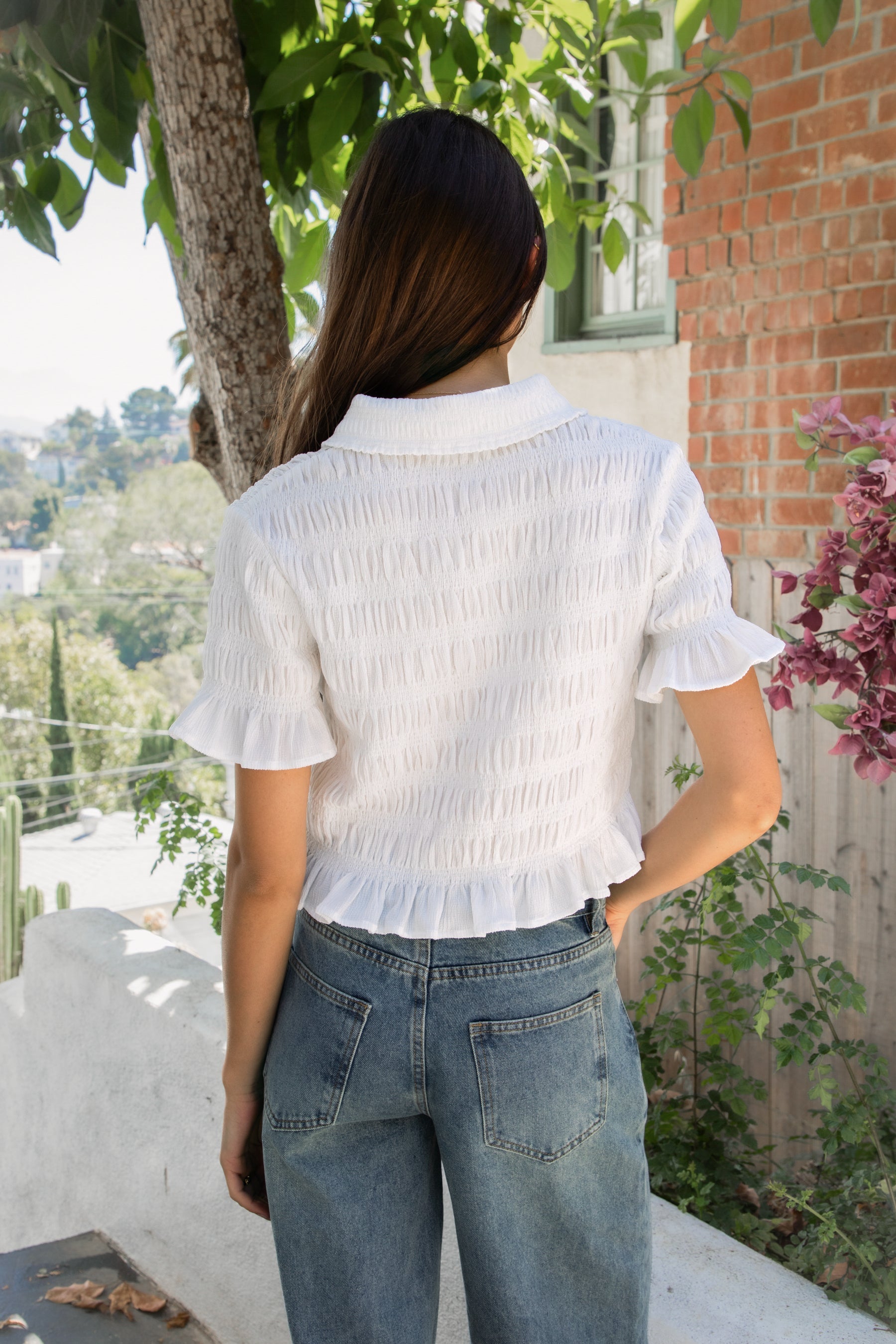 Woman wearing LISETTE smocked button front collared crop top featuring allover smocked textured fabric, soft stretch fit, classic collared neckline, functional button front closure, short puff sleeves with flounce ruffle cuffs, and a cropped silhouette with delicate ruffle hem.