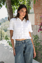 Woman wearing LISETTE smocked button front collared crop top featuring allover smocked textured fabric, soft stretch fit, classic collared neckline, functional button front closure, short puff sleeves with flounce ruffle cuffs, and a cropped silhouette with delicate ruffle hem.