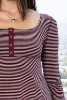 Woman wearing ELODY striped babydoll square neck henley peplum top featuring soft striped knit fabric, wide scoop neckline with contrast trim, henley inspired front placket with metal snap details, fitted bodice, empire waist seam, flared peplum hem, and long fitted sleeves.