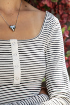 Woman wearing ELODY striped babydoll square neck henley peplum top featuring soft striped knit fabric, wide scoop neckline with contrast trim, henley inspired front placket with metal snap details, fitted bodice, empire waist seam, flared peplum hem, and long fitted sleeves.
