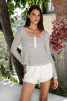 Woman wearing ELODY striped babydoll square neck henley peplum top featuring soft striped knit fabric, wide scoop neckline with contrast trim, henley inspired front placket with metal snap details, fitted bodice, empire waist seam, flared peplum hem, and long fitted sleeves.