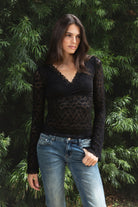 Woman wearing EMMELINE ruched lace long sleeve top featuring sheer floral stretch lace, fitted feminine silhouette, deep V neckline with scalloped lace trim, ruched detailing through the front and sides, soft mesh bust lining, sheer fitted long sleeves, flared lace trimmed cuffs, and scalloped hem finish.