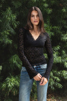 Woman wearing EMMELINE ruched lace long sleeve top featuring sheer floral stretch lace, fitted feminine silhouette, deep V neckline with scalloped lace trim, ruched detailing through the front and sides, soft mesh bust lining, sheer fitted long sleeves, flared lace trimmed cuffs, and scalloped hem finish.