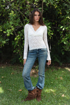 Woman wearing EMMELINE ruched lace long sleeve top featuring sheer floral stretch lace, fitted feminine silhouette, deep V neckline with scalloped lace trim, ruched detailing through the front and sides, soft mesh bust lining, sheer fitted long sleeves, flared lace trimmed cuffs, and scalloped hem finish.