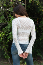 Woman wearing EMMELINE ruched lace long sleeve top featuring sheer floral stretch lace, fitted feminine silhouette, deep V neckline with scalloped lace trim, ruched detailing through the front and sides, soft mesh bust lining, sheer fitted long sleeves, flared lace trimmed cuffs, and scalloped hem finish.