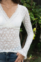 Woman wearing EMMELINE ruched lace long sleeve top featuring sheer floral stretch lace, fitted feminine silhouette, deep V neckline with scalloped lace trim, ruched detailing through the front and sides, soft mesh bust lining, sheer fitted long sleeves, flared lace trimmed cuffs, and scalloped hem finish.