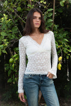 Woman wearing EMMELINE ruched lace long sleeve top featuring sheer floral stretch lace, fitted feminine silhouette, deep V neckline with scalloped lace trim, ruched detailing through the front and sides, soft mesh bust lining, sheer fitted long sleeves, flared lace trimmed cuffs, and scalloped hem finish.