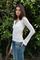 Woman wearing EMMELINE ruched lace long sleeve top featuring sheer floral stretch lace, fitted feminine silhouette, deep V neckline with scalloped lace trim, ruched detailing through the front and sides, soft mesh bust lining, sheer fitted long sleeves, flared lace trimmed cuffs, and scalloped hem finish.