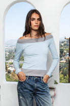 Woman wearing AVALYN contrast rib off shoulder long sleeve top featuring soft stretch knit fabric, light blue body, contrast grey ribbed trim, wide off shoulder neckline, sporty ribbed neckline, long fitted sleeves, fitted bodice, contrast ribbed hem, and hip length silhouette.