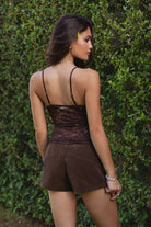 Woman in a tic toc cami top in dainty brown floral lace paired with faux suede lace up shorts, paired with frye boots, standing in front of green foliage