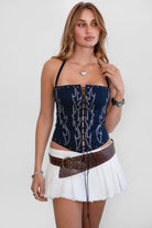 Woman wearing TIC TOC TA31346 RAELY stretch denim embroidered western corset top with halter tie and lace-up front detail with a white mini skirt and belt.