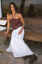 Woman wearing TIC TOC TC31322 KEZIA Mesh Sequin Embroidered Top with a matching maxi skirt and boots, sitting on a wooden bench. 