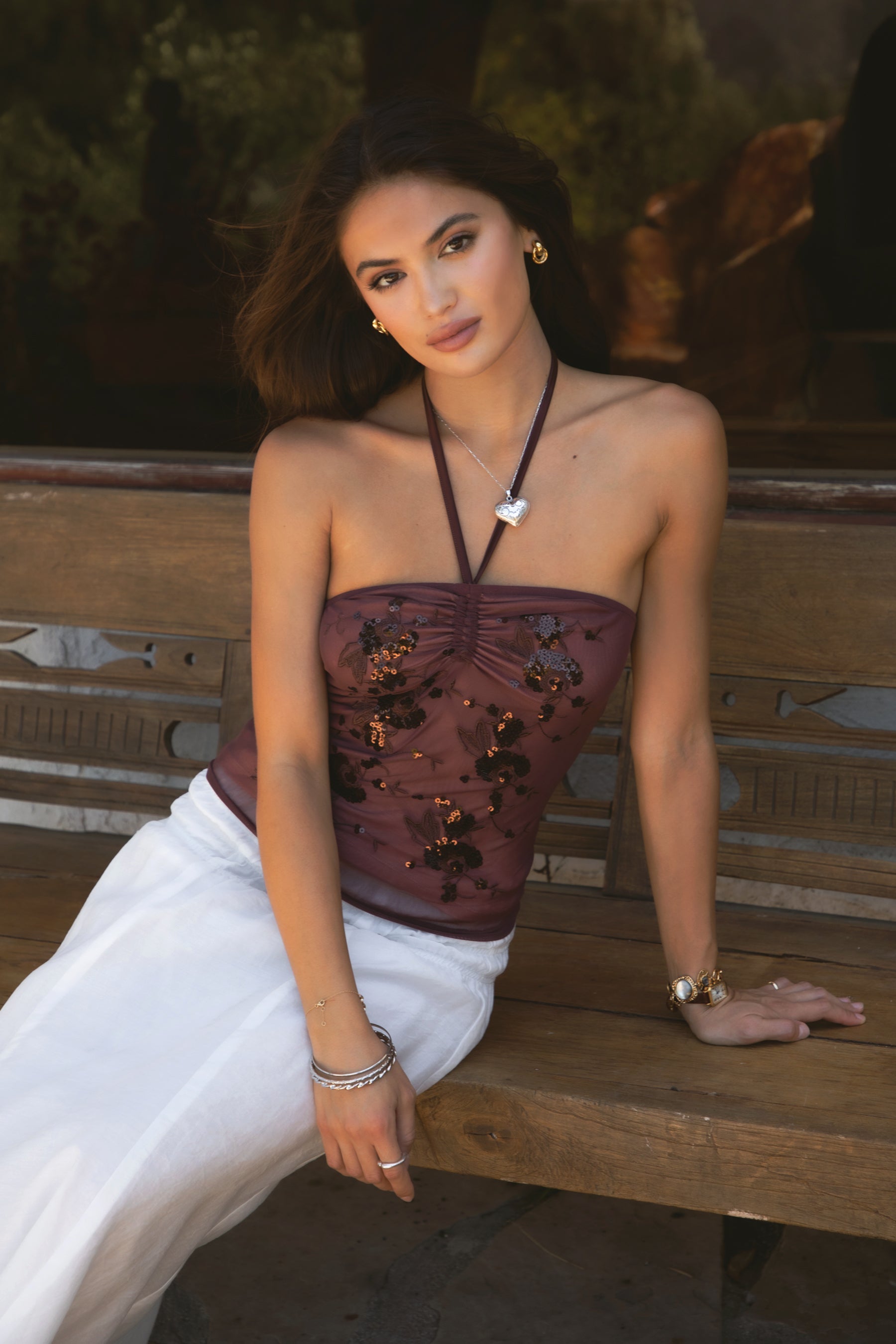 Woman wearing TIC TOC TC31322 KEZIA Mesh Sequin Embroidered Top with a matching maxi skirt and boots, sitting on a wooden bench. 