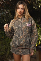 Woman wearing TIC TOC TJ31541-RODEOGIRL RODEOGIRL dark camo cloud fleece hoodie featuring an allover Bayou camo print, oversized relaxed hoodie silhouette, soft cozy cloud fleece fabric, dropped shoulder long sleeves, adjustable drawstring hood, and RODEO GIRL graphic print detail, styled with casual bottoms for a western streetwear-inspired lounge look.