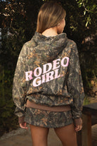 Woman wearing TIC TOC TJ31541-RODEOGIRL RODEOGIRL dark camo cloud fleece hoodie featuring an allover Bayou camo print, oversized relaxed hoodie silhouette, soft cozy cloud fleece fabric, dropped shoulder long sleeves, adjustable drawstring hood, and RODEO GIRL graphic print detail, styled with casual bottoms for a western streetwear-inspired lounge look.