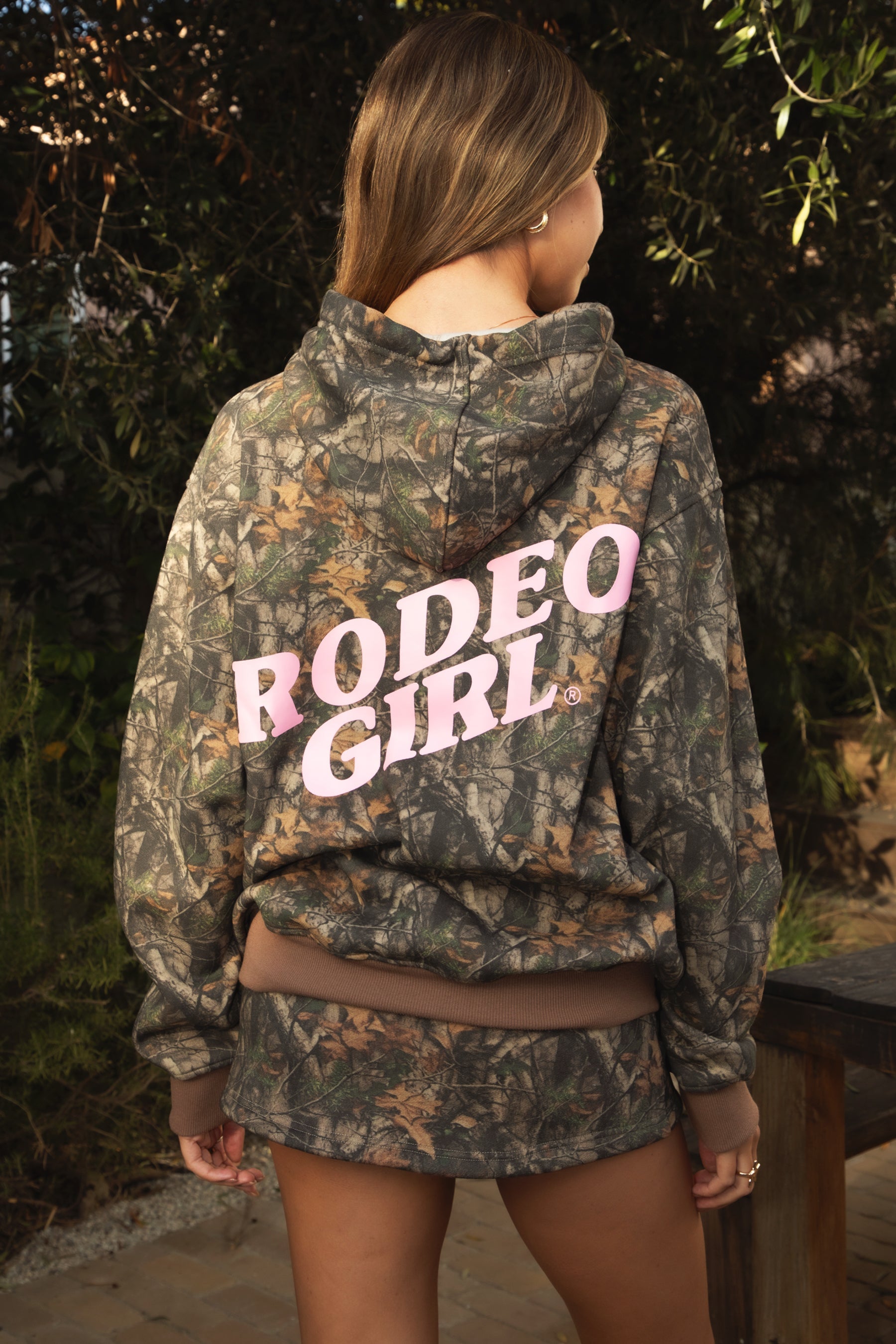 Woman wearing TIC TOC TJ31541-RODEOGIRL RODEOGIRL dark camo cloud fleece hoodie featuring an allover Bayou camo print, oversized relaxed hoodie silhouette, soft cozy cloud fleece fabric, dropped shoulder long sleeves, adjustable drawstring hood, and RODEO GIRL graphic print detail, styled with casual bottoms for a western streetwear-inspired lounge look.