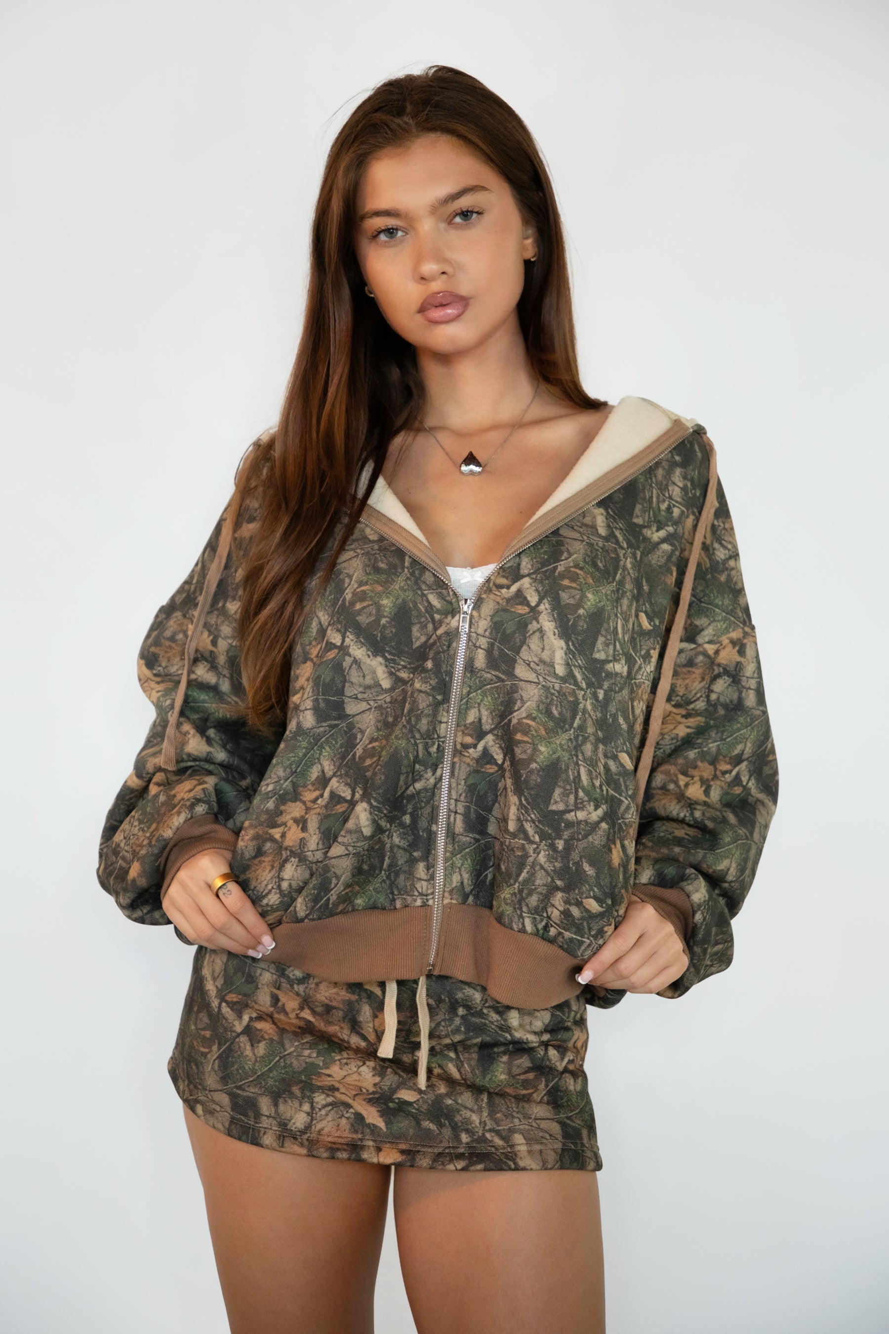 Woman wearing TIC TOC TJ31823-BAYOU-JK BAYOU dark woodland camo fleece zip up hoodie featuring an oversized relaxed fit, full zip front closure, attached drawstring hood, front kangaroo pocket, and ribbed cuffs and hem, styled with casual bottoms for a cozy everyday lounge-and-street look.