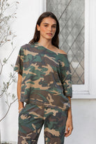 Woman wearing SCOUT military camo short sleeve oversized off shoulder top featuring soft breathable single jersey fabric, allover classic camouflage print, wide rolled raw edge neckline, relaxed oversized silhouette, dropped shoulders, elbow length sleeves, and casual boyfriend inspired draped fit.