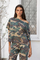 Woman wearing SCOUT military camo short sleeve oversized off shoulder top featuring soft breathable single jersey fabric, allover classic camouflage print, wide rolled raw edge neckline, relaxed oversized silhouette, dropped shoulders, elbow length sleeves, and casual boyfriend inspired draped fit.