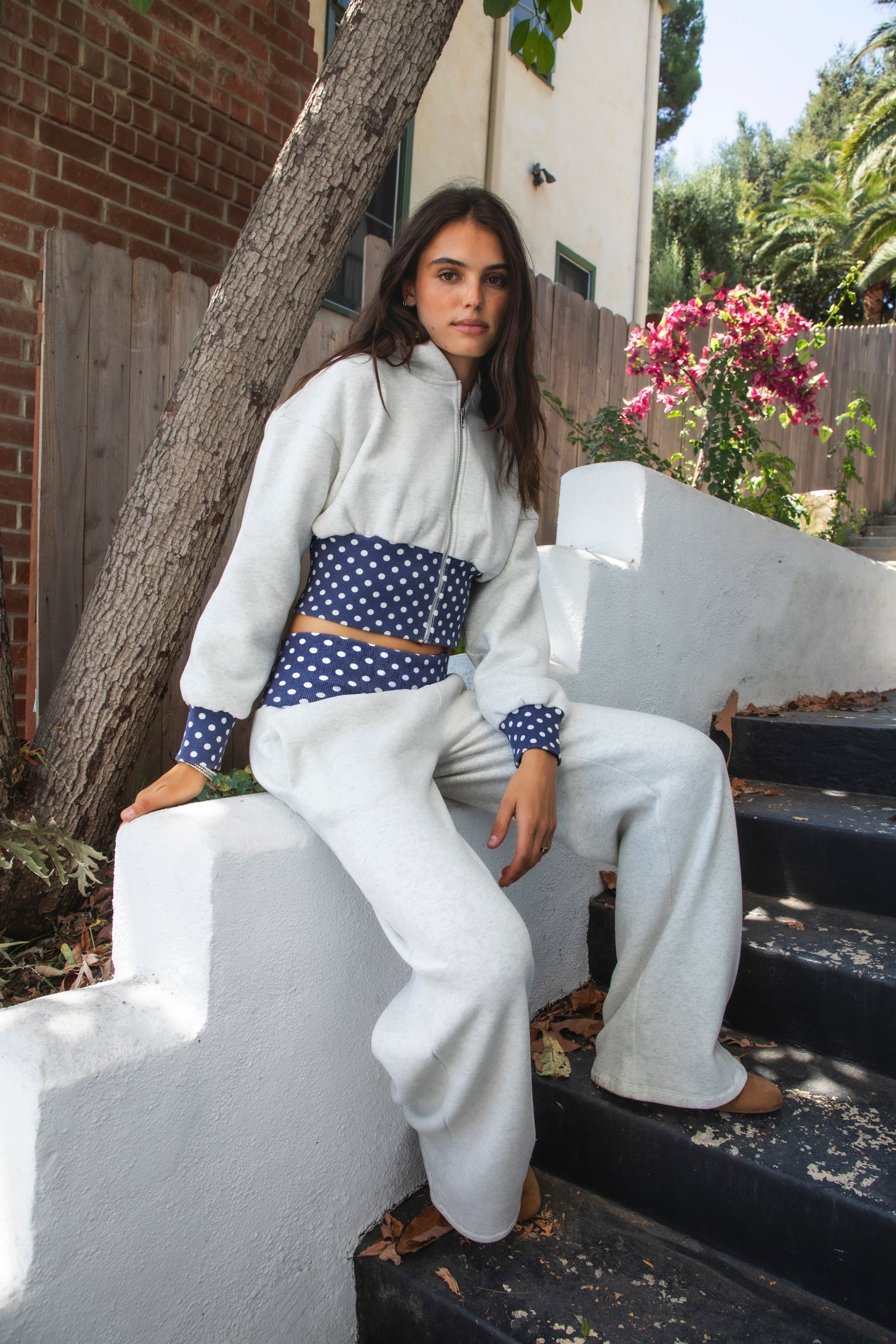 Woman wearing COSETTE polka dot corset waist cropped zip up hoodie featuring soft fleece fabric, contrast navy ribbed corset inspired waistband with allover white polka dot pattern, fitted extended waist, matching polka dot cuffs, full front zipper closure, relaxed drop shoulder sleeves, attached hood, and cropped length designed to coordinate with the matching pants.