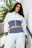 Woman wearing COSETTE polka dot corset waist cropped zip up hoodie featuring soft fleece fabric, contrast navy ribbed corset inspired waistband with allover white polka dot pattern, fitted extended waist, matching polka dot cuffs, full front zipper closure, relaxed drop shoulder sleeves, attached hood, and cropped length designed to coordinate with the matching pants.