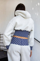 Woman wearing COSETTE polka dot corset waist cropped zip up hoodie featuring soft fleece fabric, contrast navy ribbed corset inspired waistband with allover white polka dot pattern, fitted extended waist, matching polka dot cuffs, full front zipper closure, relaxed drop shoulder sleeves, attached hood, and cropped length designed to coordinate with the matching pants.