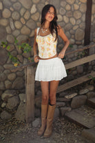 Woman wearing TIC TOC ELORA Floral Lace Bustier Top, front view showing the sweetheart neckline and lace shoulder straps, paired with a white tiered mini skirt, and tan boots outdoors. 