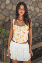 Woman wearing TIC TOC ELORA Floral Lace Bustier Top, front view showing the sweetheart neckline and lace shoulder straps, paired with a white tiered mini skirt, outdoors. 